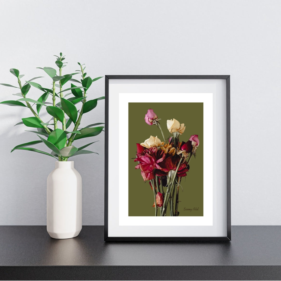 Framed artwork of various flowers in a vase on a green background, displayed on a black shelf next to a potted plant in a white vase.