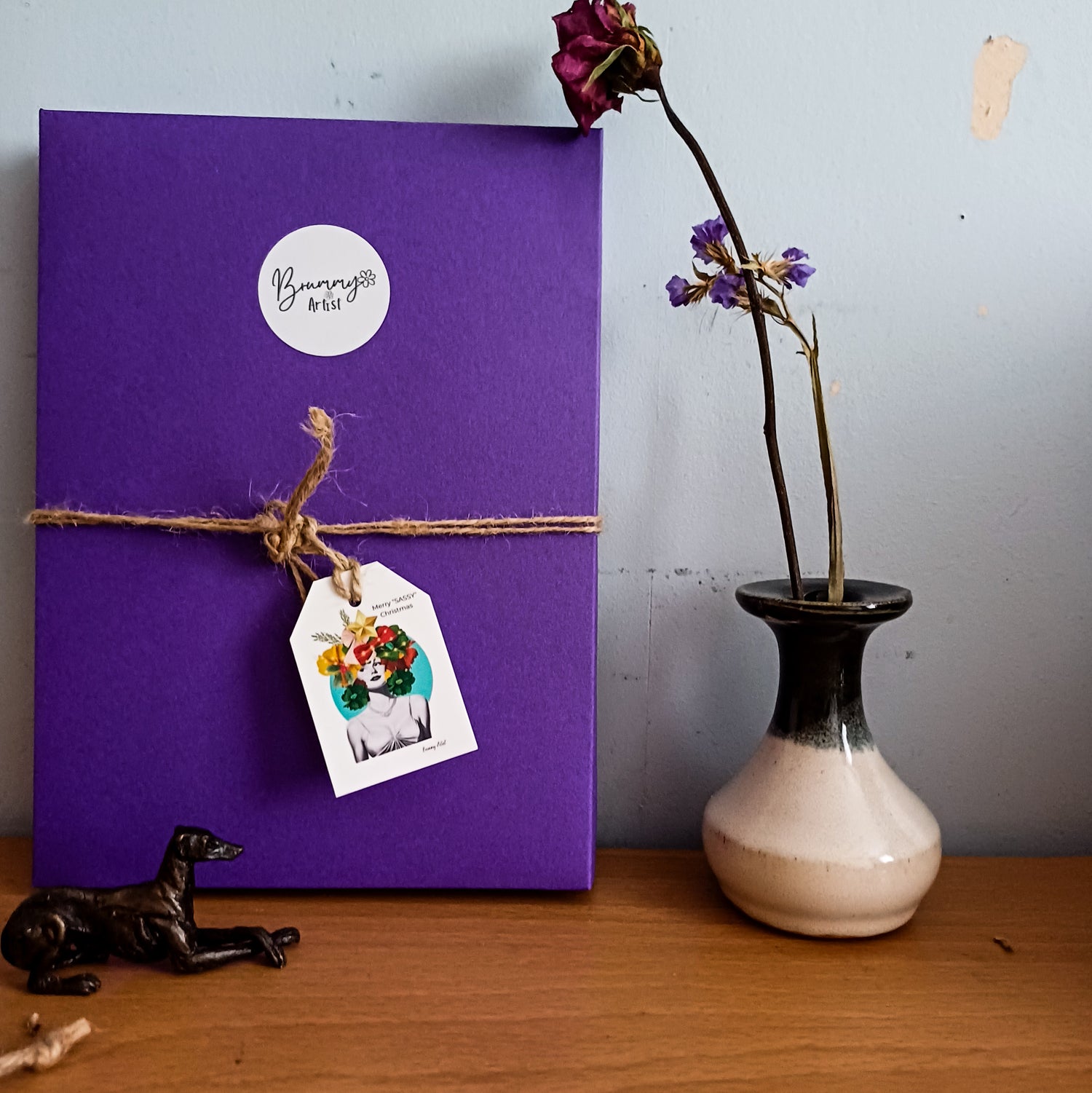 The Luxury Gift wrapped in a purple box, featuring a twine bow and flower tag from the brand Wrapped, adds a special touch beside a ceramic vase with dried flowers. A small black dog figurine completes the charming scene on the wooden surface.