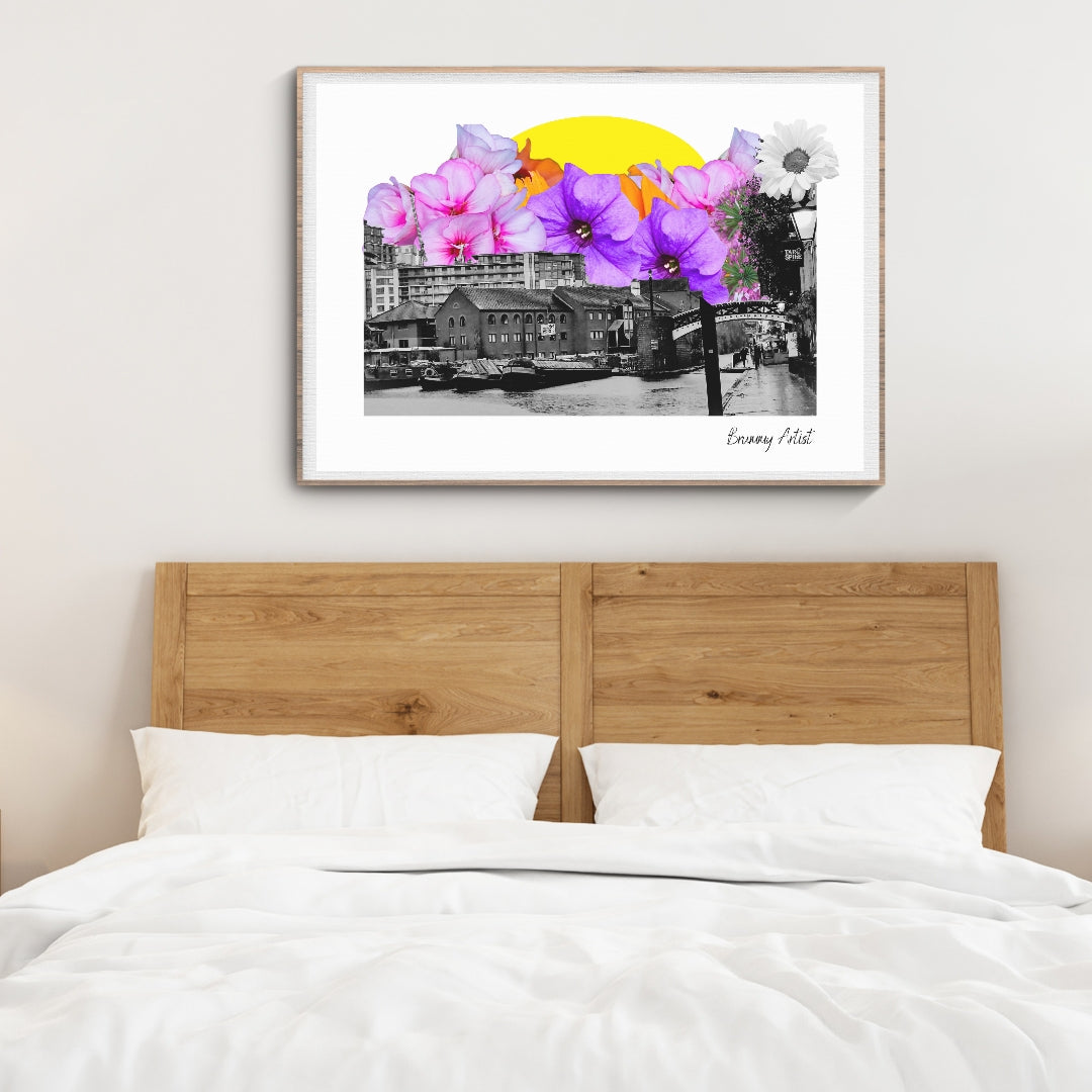 A wooden bed with white bedding sits below a framed art print from The Brummy Artist UK, titled "Brumside Blooms - Brindley Place Birmingham," featuring a floral collage over an urban cityscape enhanced by a yellow circle.