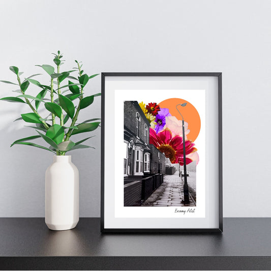 On a shelf sits a framed piece titled "A Moment in Time in Aston Birmingham - floral collage art print" by The Brummy Artist UK, showcasing vibrant collage art of a black and white street scene, enriched with colorful homegrown flora and an orange sun, alongside a plant.